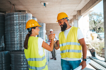 A smiling male and female construction worker shake hands on a busy construction site, attired in safety vests and hard hats, reflecting teamwork and collaboration in a professional setting.