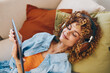 © SHOTPRIME STUDIO - Happy Woman Holding Tablet PC and Headphones, Enjoying Music and Chatting Online on Couch in Cozy Living Room