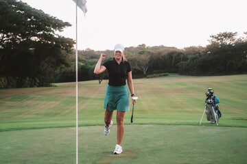 A professional golfer, a young woman in a sports skirt and cap, hits the hole with the ball. She raised her hand up and rejoiced at her victory, opening her mouth and smiling. Hitting the target.