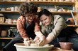 © AnnaStills - Low angle view shot of two diverse women wearing aprons having fun during pottery class shaping clay on wheel together