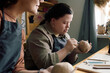 © AnnaStills - Selective focus shot of young woman with Down syndrome using clay carving tools and chatting with her friend during pottery class
