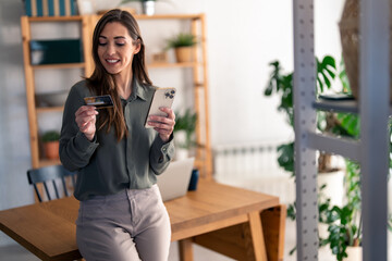 Happy young woman customer shopper holding credit card using cell phone mobile app paying online making purchase in e-commerce digital store on smartphone at home.