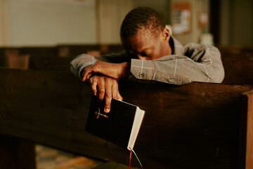 Selective focus shot of unrecognizable African American man sitting on pew in Catholic church holding Bible book and praying to God