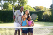 © wavebreak3 - Outdoors, diverse family of four is smiling and holding tennis rackets on sunny day