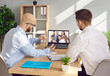 © Studio Romantic - Team of business people meeting online via video call. Two young men sitting together at office desk, looking at laptop computer screen and talking to diverse group of remote international colleagues