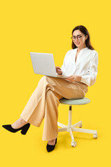 Wall Mural - Pretty young woman working with laptop while sitting on chair against yellow background