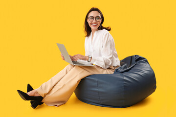 Wall Mural - Pretty young woman working with laptop while sitting on bean bag chair against yellow background
