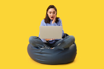 Wall Mural - Surprised young woman with laptop sitting on bean bag chair against yellow background