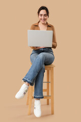 Wall Mural - Pretty young woman with laptop sitting on chair against beige background