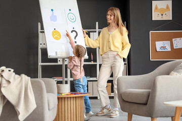 Wall Mural - Little girl with psychologist pointing at thumb-up on board in office