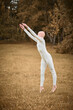 © TRAVELARIUM - Full length portrait of young hairless girl ballerina with alopecia in tight white suit prepares to jump on fall lawn in park, symbolizing overcoming challenges and gracefully acceptance individuality