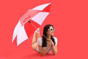 Beautiful young woman in beachwear with umbrella lying on red background