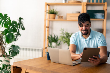 Handsome male entrepreneur using digital tablet sitting at the table in a home office, looking at the paper, communicating online, writing emails, distantly working or studying on computer at home.