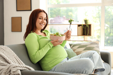 Young pregnant woman with fresh vegetable salad sitting on sofa at home