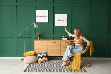 Sticker - Young woman in armchair with hanging posters on green wall at home