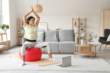 Poster - Sporty African-American woman with laptop practising yoga on fitball at home