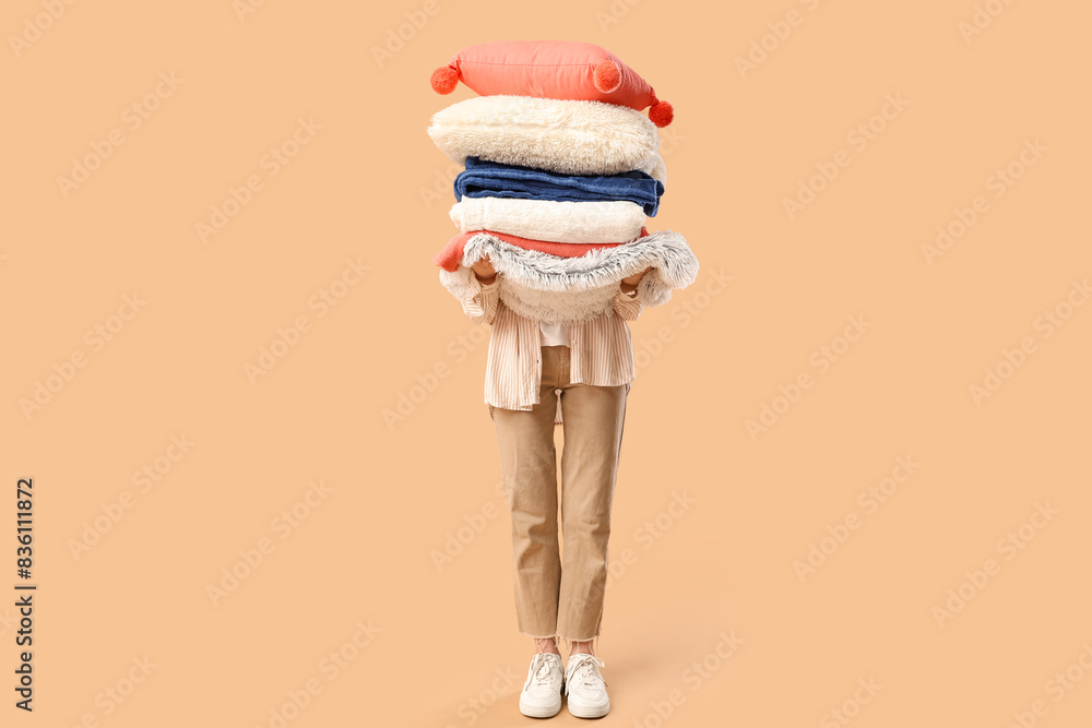 Beautiful young woman with stack of different blankets and pillows on beige background
