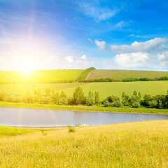 Wall Mural - Summer landscape with a green field and a lake. Sky with bright sun and clouds.