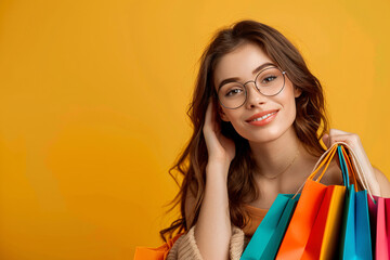 Wall Mural - Cheerful young woman in glasses holding shopping bags over a vibrant yellow backdrop