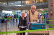 © luciano - Couple of senior travelers with suitcases pushing luggage trolley while walking through airport area to reach check in area and departure gate. Old senior man and woman leaving for vacation