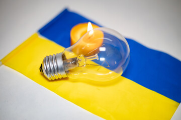 Wall Mural - Light bulb on the background of a burning candle and the Ukrainian flag. Blackout due to the war in Ukraine