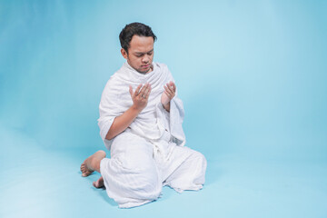 Serious young Asian Muslim man wearing ihram sitting down and praying with hand gesture over isolated by blue background. Hajj concept.