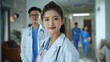 © MiguelAngel - Young Asian female doctor, posing looking at the camera, with doctors out of focus in the background, inside the hospital.