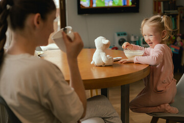 Baby girl eats breakfast and watches cartoons on TV with mother. Children and technology concept. Alpha Generation and modern technology with electronic device. Part of the series