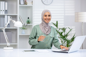 Wall Mural - Confident businesswoman wearing hijab sitting at desk with laptop and glass of water, working in modern office. Professional career and productivity concept.