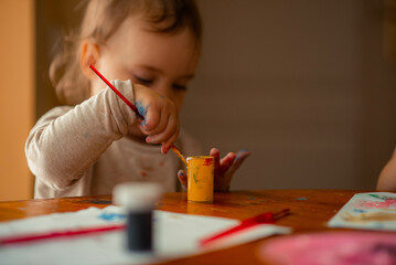 Wall Mural - a little girl of two years old sits at the table and paints with a brush, she stains her hands with paint and smiles