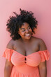 © sommersby - A woman with curly hair smiles against a pink wall. She is wearing a peach off-the-shoulder dress