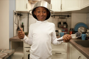 Poster - Portrait of black boy kid in blank mockup long-sleeve standing with cooking pan on head and whisk in hands, ready to help his mom to cook delicious dinner or bake cake for his birthday party