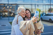 © Dragana Gordic - A joyful senior mother and her adult daughter pose for a selfie, phone in hand, outside a grocery store, bags of shopping in tow, showcasing intergenerational bonding.