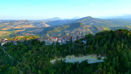 Wall Mural - Pristine aerial pullback shot from Penna San Giovanni (MC) with the flourishing thickets perched on a cliff, surrounding the town centre with its old-styled buildings, Mount Ascensione in the distance