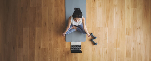 High angle view of young Asian woman practicing yoga and meditation at home sitting on floor in living room in lotus position with laptop and dumbbell. Mindful meditation and wellbeing concept