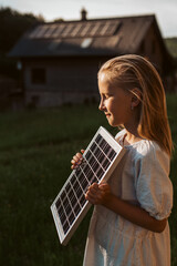 Wall Mural - Little girl with model of solar panel, standing in the middle of meadow, house with solar panels behind. Concept of renewable resources.