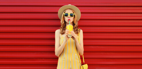 Wall Mural - Beautiful stylish young woman drinking fresh juice holding cup wearing summer hat on red background