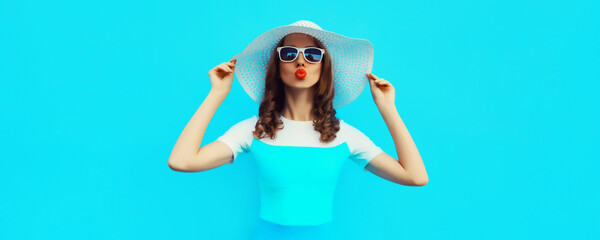 Wall Mural - Portrait of beautiful young woman posing blowing kiss wearing summer straw hat on blue background