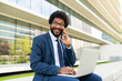 © Westend61 - Smiling businessman sitting with laptop and talking on smart phone in front of building