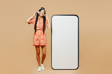 Wall Mural - Full body young woman wears orange dress casual clothes big huge blank screen mobile cell phone smartphone with area listen to music in headphones isolated on plain beige background Lifestyle concept