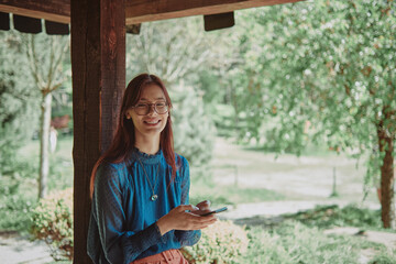 Wall Mural - Joyful teenage girl using smartphone in a rustic wooden gazebo, surrounded by a lush garden. Cheerful lifestyle portrait highlighting digital engagement and outdoor relaxation.