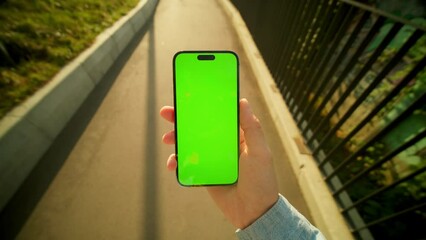 Shooting of man looking at smartphone with green screen while walking on bridge. Guy reading message on phone on blurred background of nature. Outside. Daytime