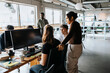 © Maskot - Young female entrepreneur standing by colleagues discussing while sitting on chair at office
