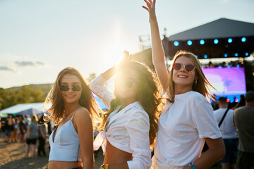 Three happy female friends dance at sunny beach music fest. Crowd enjoys summer vibes near stage. Girls in casual wear celebrate, have fun in festival atmosphere by sea shore during daytime.