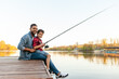 © Богдан Маліцький - African American man and child sitting on a wooden pier holding fishing rods on the river, dad teaching his son to fish in the lake, family resting and relaxing on the weekend