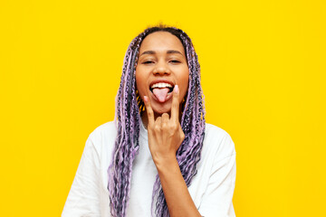 cheerful african american woman with colored dreadlocks shows rock and tongue gesture on yellow isolated background, crazy hipster girl with purple braids and unique hairstyle shows heavy music sign