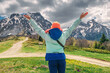© EdNurg - A woman in a jacket and orange hat is standing in a field with mountains in the background. She is smiling and enjoying the beautiful scenery