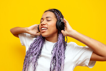 Wall Mural - cheerful african american woman with colored dreadlocks listens to music on headphones on yellow isolated background, hipster girl with purple braids and unique hairstyle listens to songs