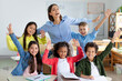 © Home-stock - Excited diverse schoolchildren raising arms and smiling, sitting in classroom with their happy young female teacher, back to school concept