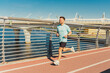 © muse studio - A man jogs on a red pathway along a waterfront, under a clear sky, with a bridge in the background.